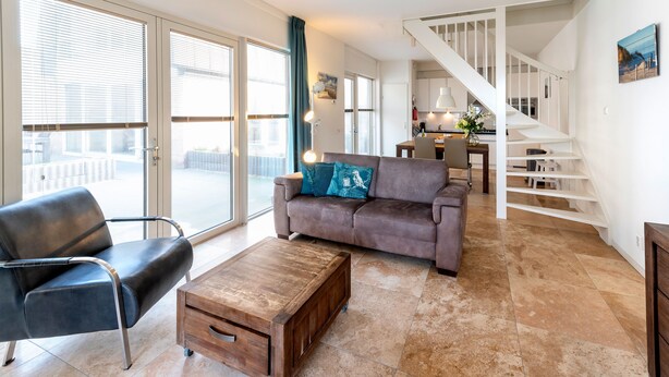 A spacious living area with a brown sofa, a wooden coffee table, and a black chair. A staircase leads to an upper floor, and a dining area is visible in the background. - Flat Fish (Egmond aan Zee)
