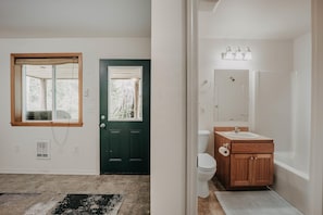 Une pièce avec une porte en bois, une fenêtre avec un rideau et une salle de bain avec un meuble-lavabo en bois et un miroir.