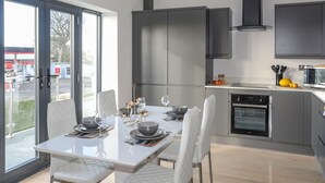 A modern kitchen with grey cabinets, a built-in oven, and a dining area with a white table and chairs. - Bryn Min Manton Apartment (Caernarfon)