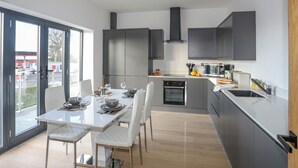 A modern kitchen with grey cabinets, a built-in oven, and a dining area with a white table and chairs. - Bryn Min Manton Apartment (Caernarfon)