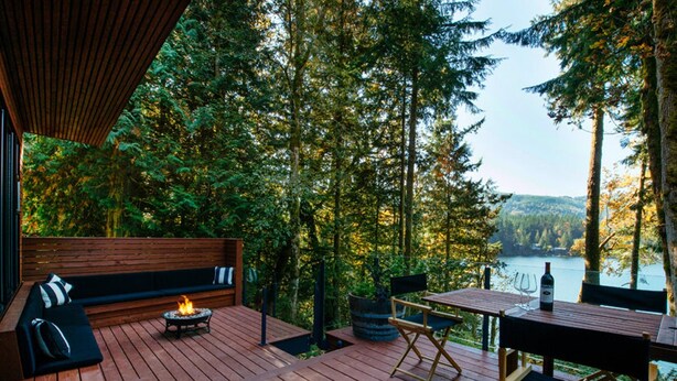 A wooden deck with a fire pit, seating, and a table with a bottle of wine, overlooking a forest and a lake. - Silver Lake 83 - Spectacular Dream Home Overlooking Silver Lake (Deming)