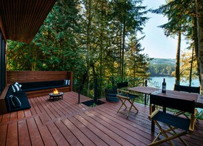 Relax on the deck overlooking a serene forest and water, surrounded by a sofa, fire pit, and table.