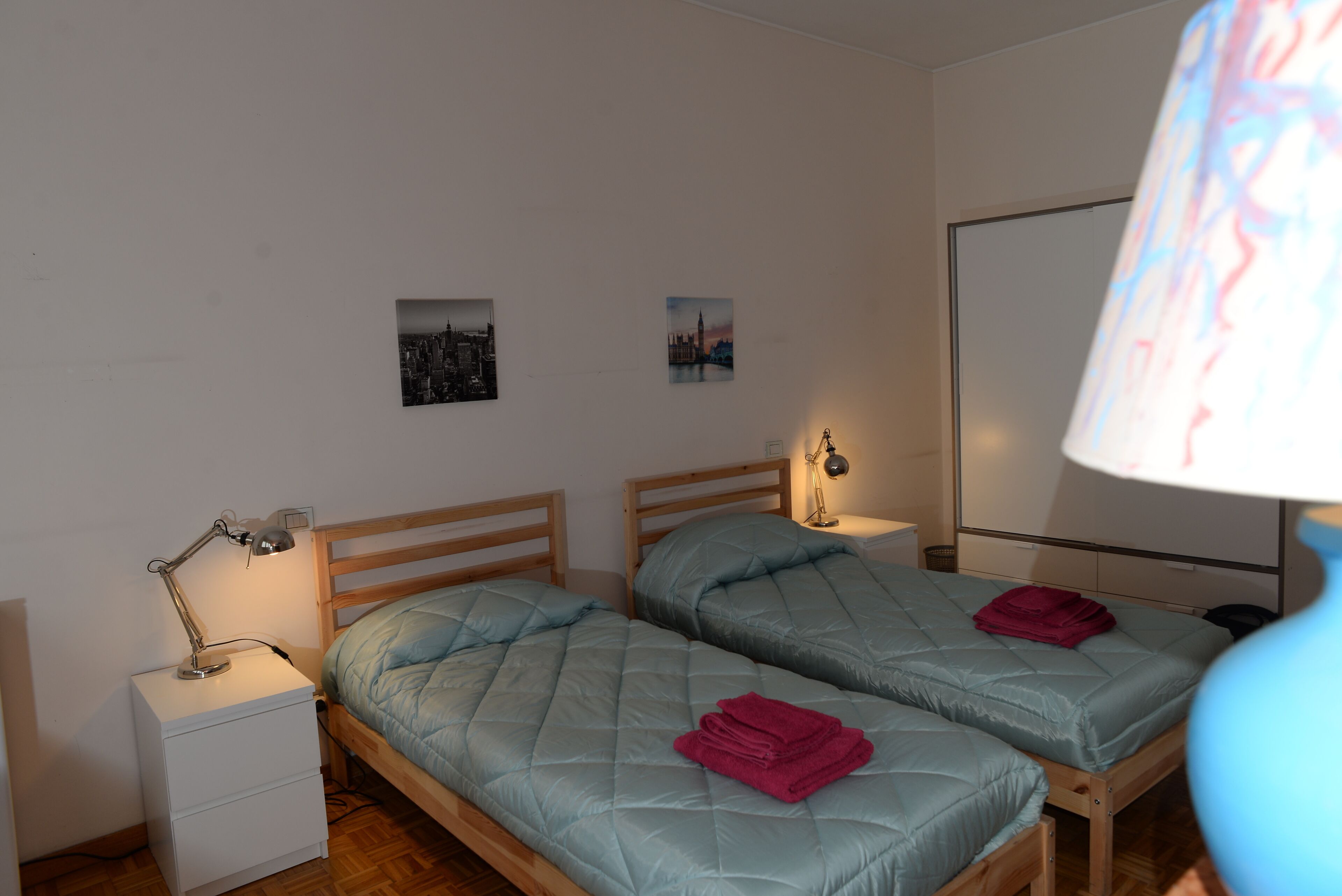 A room with two beds, a nightstand with a lamp, and a wall-mounted TV.
