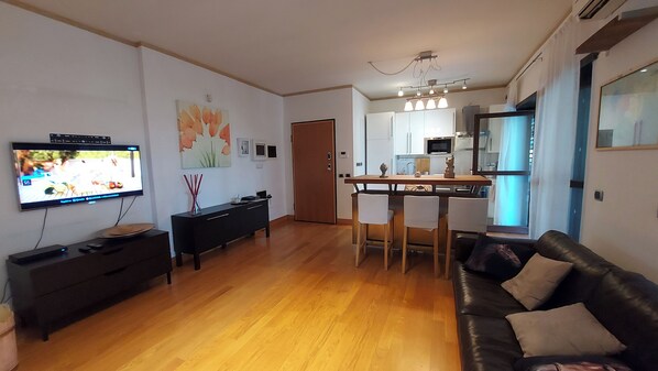 A modern living room with a flat-screen TV, a black entertainment unit, a wooden dining table with chairs, a black leather sofa, and a kitchen area with a microwave and cabinets.