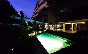 A swimming pool with a green-lit edge, surrounded by greenery and modern buildings.
