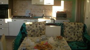 A compact kitchen with a microwave, stove, and sink. There is a dining area with a table set for two, featuring a floral tablecloth and a plate of food. - Room in Guest Room - S'olivariu Village Affittacamere - King Room With Roll-in Shower (Piscinas)