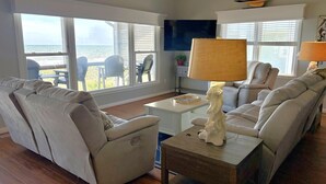 Ett vardagsrum med en soffa, fåtöljer, ett soffbord och en lampa. På väggen hänger en tv och ovanför den finns en dekoration i form av en segelbåt.