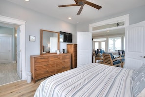 A bedroom with a bed, wooden dresser, mirror, ceiling fan, and a view of a balcony with outdoor seating.