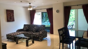 A modern living room with a sofa, a coffee table, a dining table with chairs, a ceiling fan, and large windows with curtains. - Nordic Park Hill Residence Flat (Pattaya)