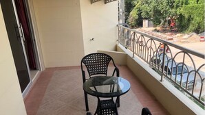 A balcony with a glass table and two wicker chairs, a metal railing, and a view of parked cars and greenery. - Nordic Park Hill Residence Flat (Pattaya)