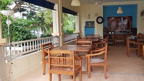 Un área de comedor al aire libre con mesas y sillas de madera, una pared azul con un letrero de comida y bebida, y un reloj en la pared.