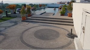 A rooftop pool area with steps leading down to the water, surrounded by potted plants and overlooking a cityscape.