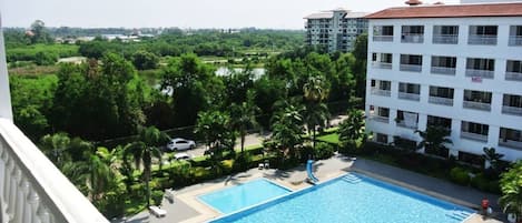 A swimming pool with a slide, surrounded by greenery and adjacent to a white building with balconies.