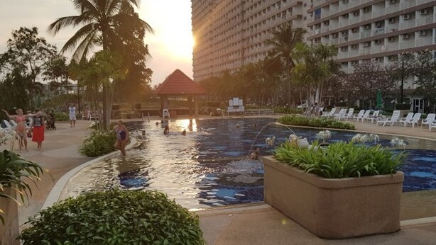 A swimming pool area with lounge chairs, palm trees, and a building in the background. - Jomtien Beach Condominium Jbc A2 F4 R28 (Pattaya)