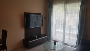 A living room with a flat-screen TV mounted on a dark wooden cabinet, a glass coffee table with a vase of flowers, and a view through a window with light blue curtains.