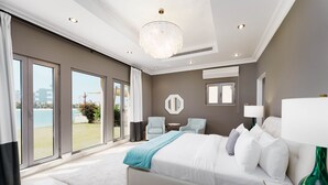 A modern bedroom with a large bed, two armchairs, a chandelier, and a view of the sea. - Maison Privee - Palm Jumeirah Beach Front XL Villa w/Prvt Pool (Dubai)