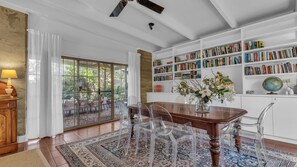 Une pièce spacieuse avec une grande table à manger en bois, des chaises transparentes, une bibliothèque remplie de livres et, par une porte vitrée, une vue sur un espace extérieur.