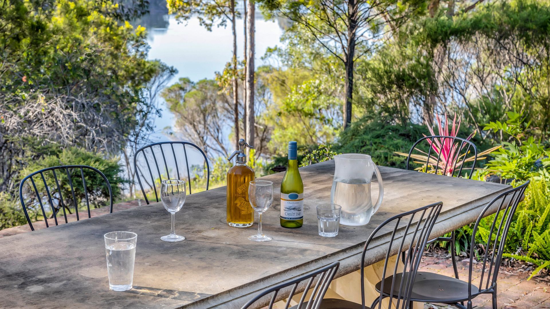 An outdoor dining table with chairs, a wine bottle, glasses, and a pitcher, set against a backdrop of trees and a body of water.