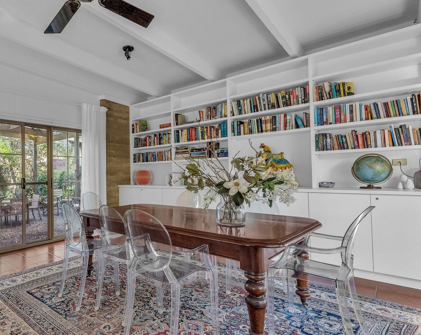 A dining area with a wooden table, clear chairs, a patterned rug, and a bookshelf filled with books.
