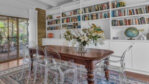 Un coin repas avec une table en bois, des chaises transparentes, un tapis à motifs et une bibliothèque remplie de livres.