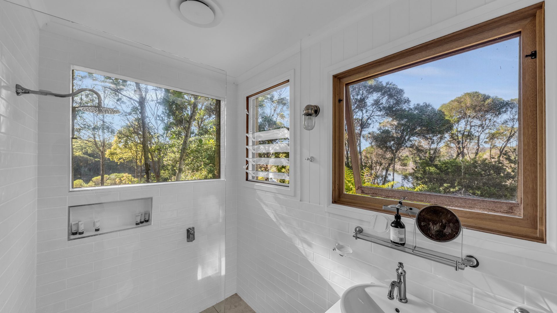 A bathroom with white tiled walls, a bathtub, a sink, and two windows offering views of trees and a lake.