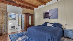 Slaapkamer met een groot bed, een houten garderobe, een badkamer met een witte wastafel en uitzicht door een geopende deur.