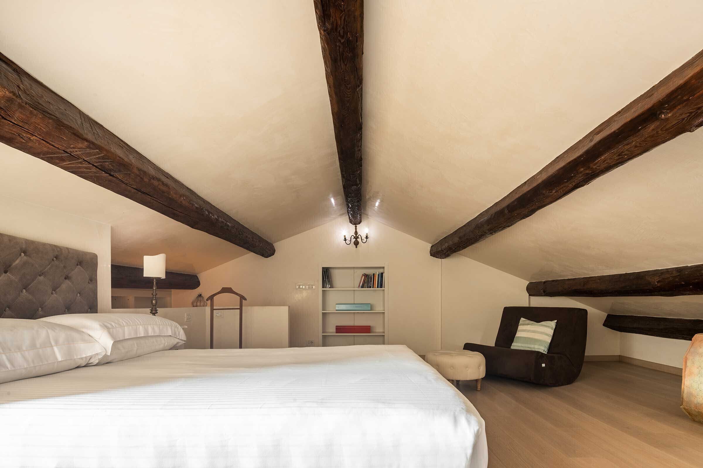 A bedroom with a large bed, a bedside table, a chair, a bookshelf, and exposed wooden beams on the ceiling.