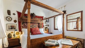 Uma cama de dossel tradicional com colcha estampada em vermelho e branco, uma mesinha de cabeceira de madeira com uma estátua de Buda e uma mesa de madeira com livros.
