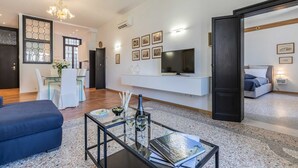A modern living room with a glass coffee table, a sofa, a dining area with a white table and chairs, a TV mounted on the wall, and a bedroom visible through an open door. - San Marco Alluring and Spacious Apartment with Elevator (Venecia)