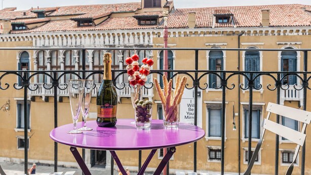 Un balcón con una mesa morada dispuesta con una botella, vasos y aperitivos.