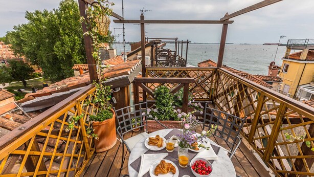 Terraço com mesa posta para o café da manhã, com vista para um corpo d'água e a paisagem urbana.