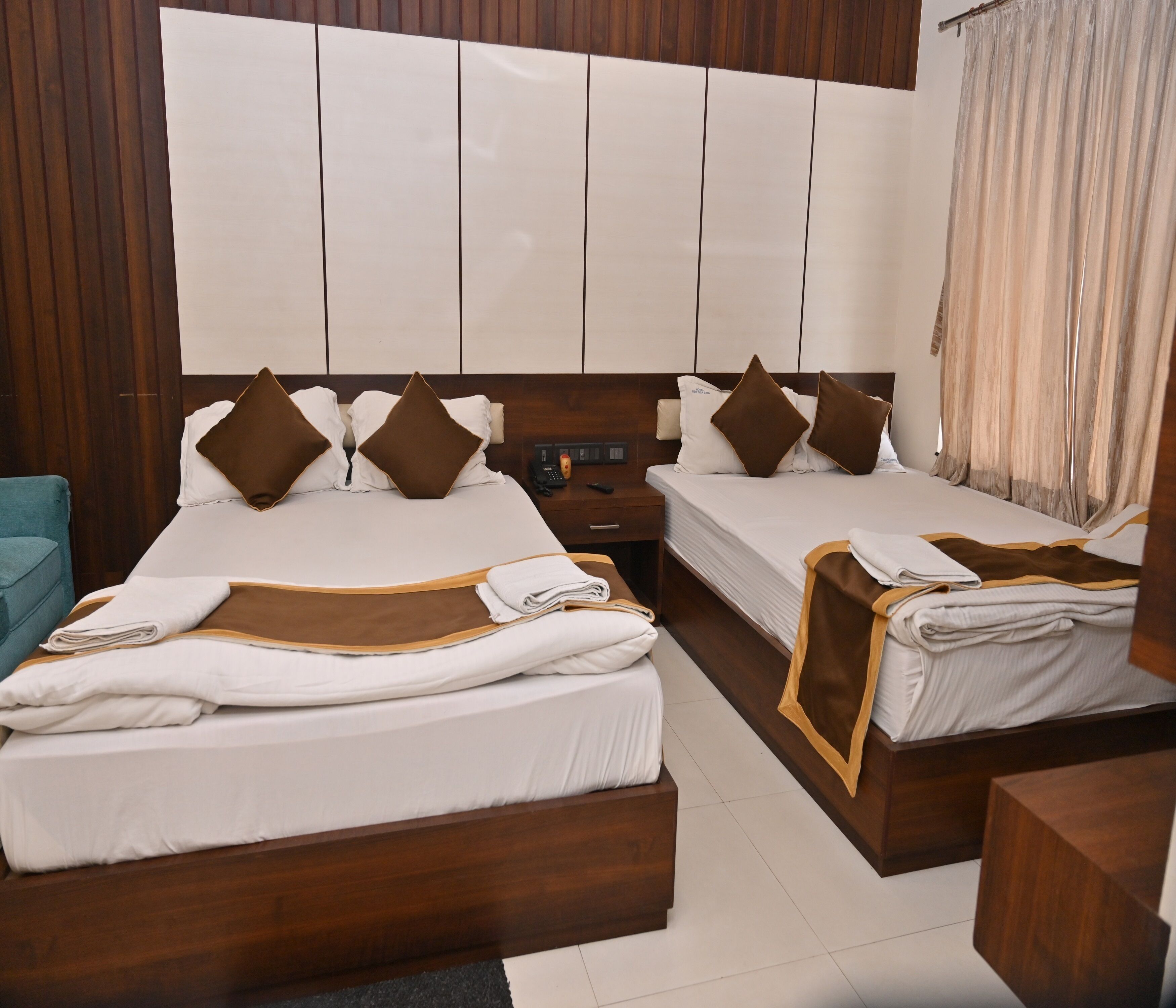 A hotel room with two beds, each with a wooden headboard, white bedding, and brown pillows. There is a wooden nightstand with a phone and a remote control, and a teal chair.