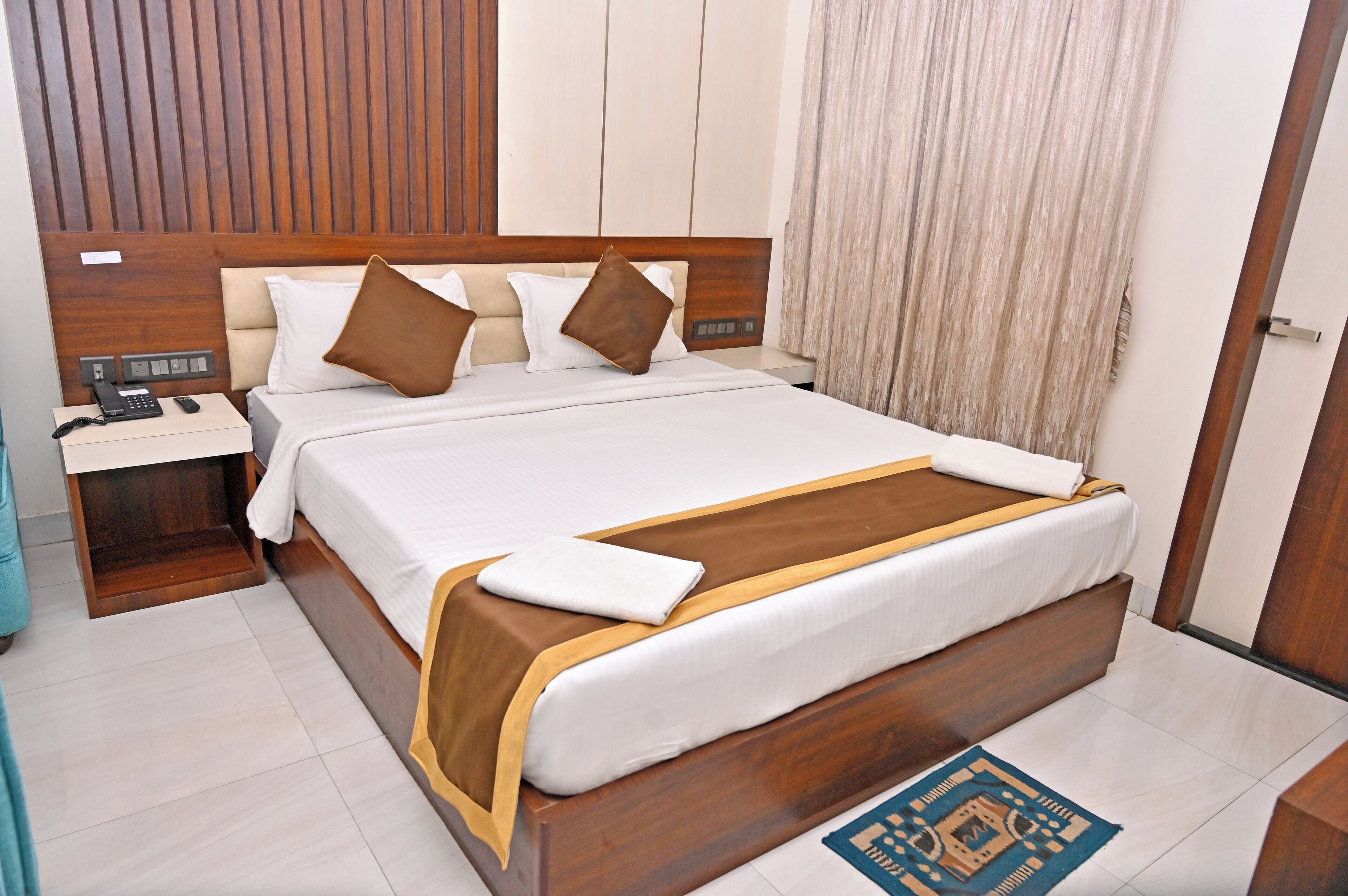 A neatly made bed with a wooden headboard, a bedside table with a phone and a lamp, and a patterned rug on the floor.