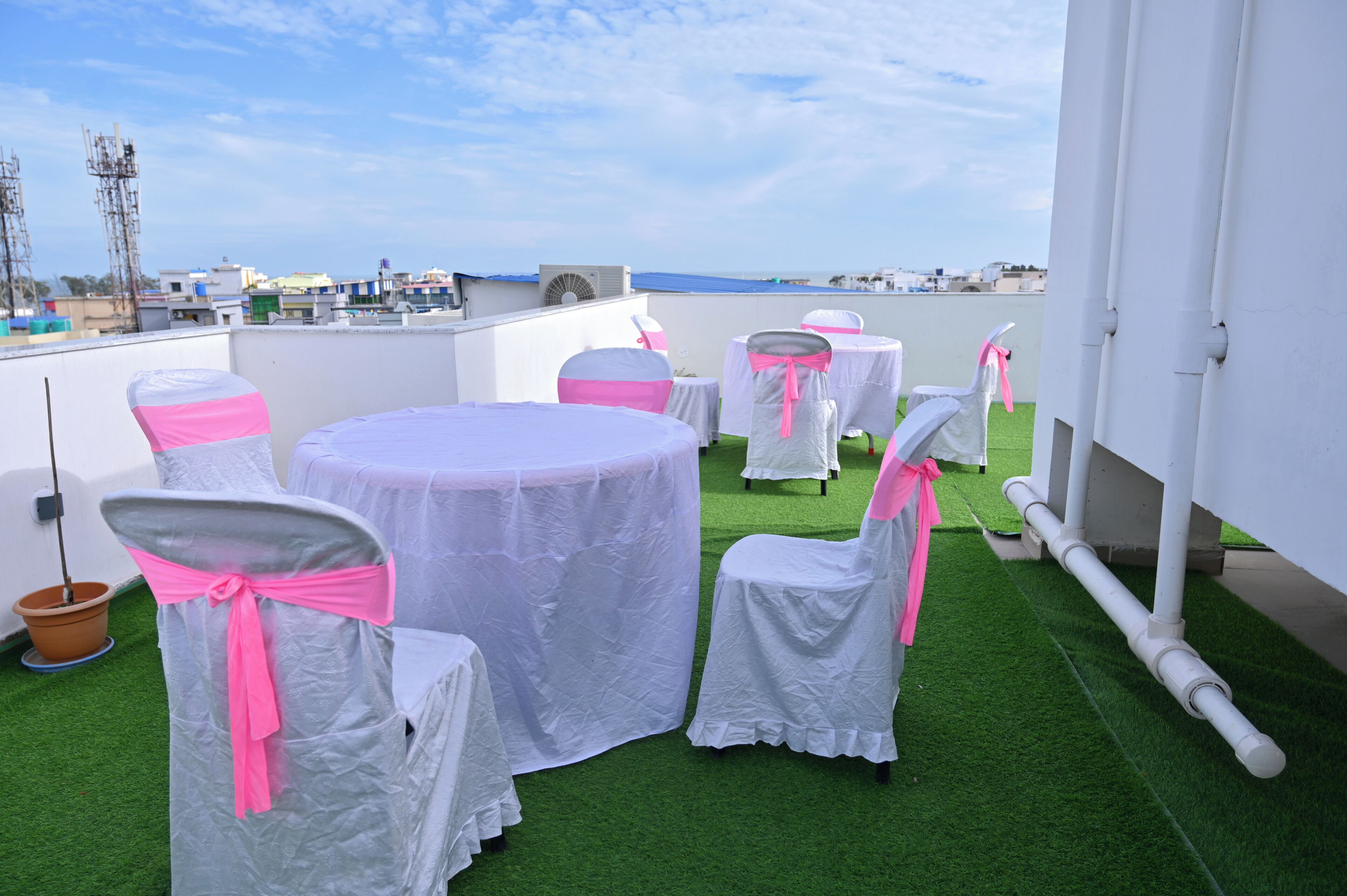 A rooftop terrace with tables and chairs covered in white tablecloths and pink chair sashes, set on artificial green turf.