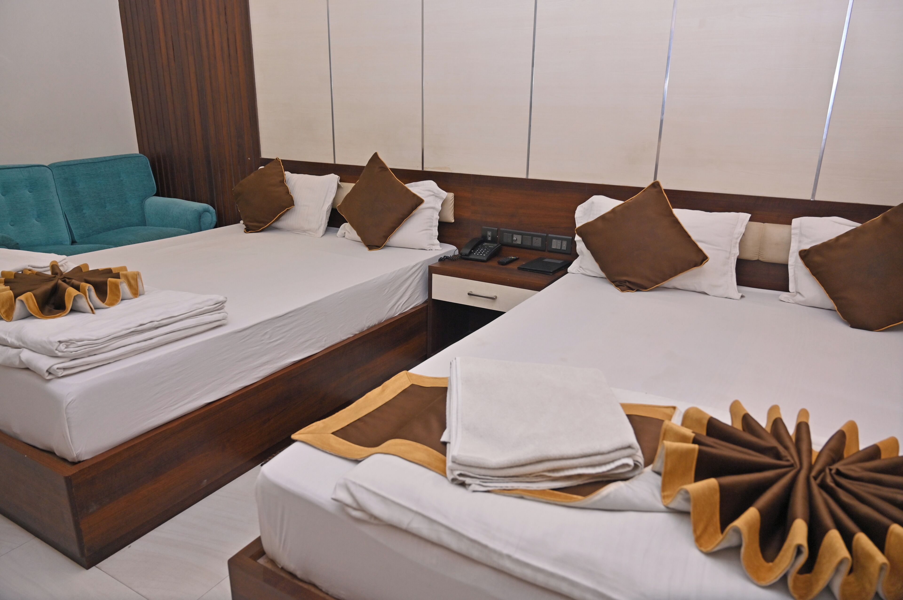A hotel room with two beds, a wooden headboard, a nightstand with a phone and a book, and a teal sofa.