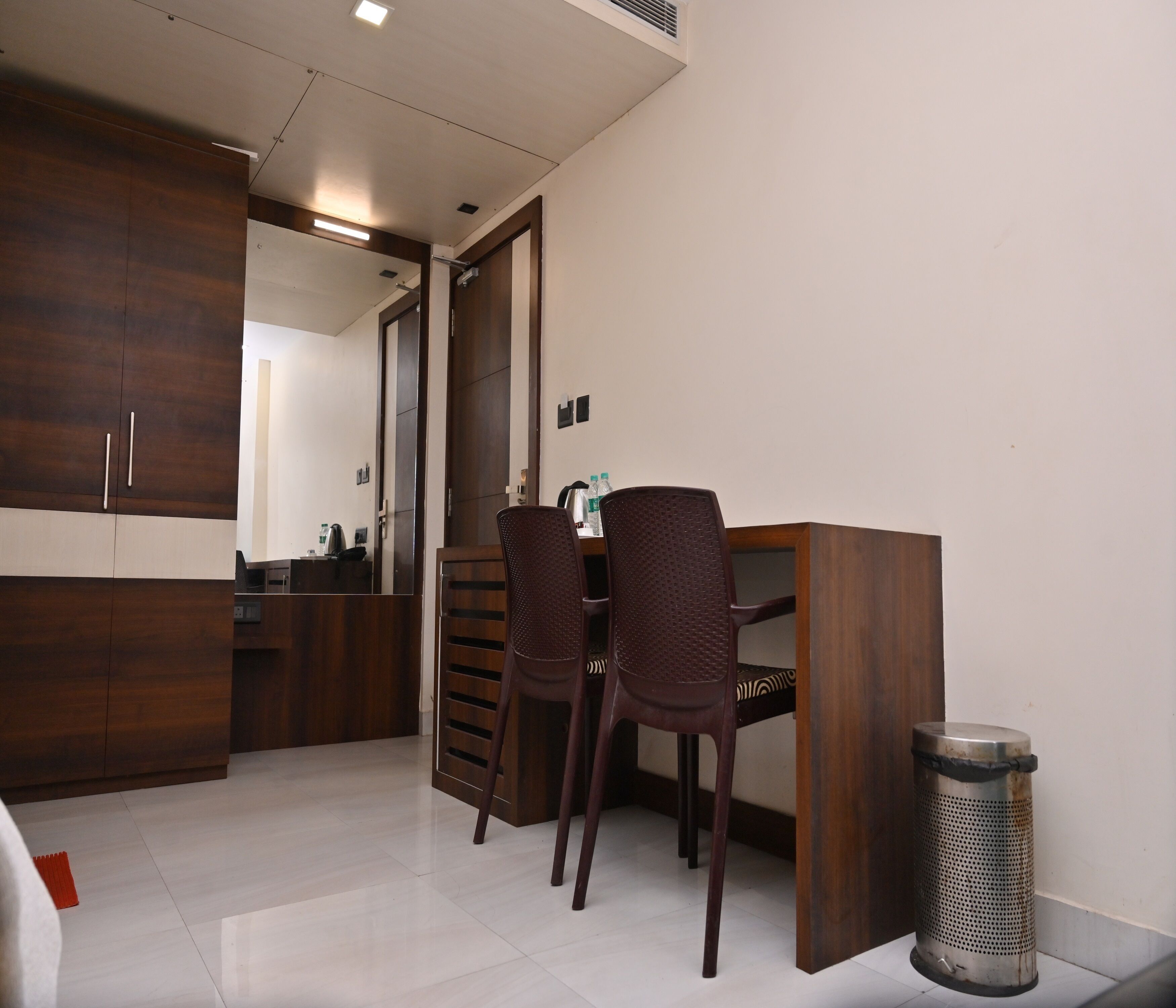 Una habitación de hotel con un armario de madera, un escritorio con dos sillas, un cubo de basura y un espejo.