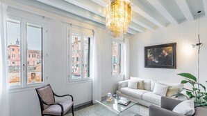 Un salon moderne avec un canapé blanc, une table basse en verre et un fauteuil. De grandes fenêtres sont ornées de rideaux, un lustre illumine la pièce et un tableau encadré est accroché au mur.