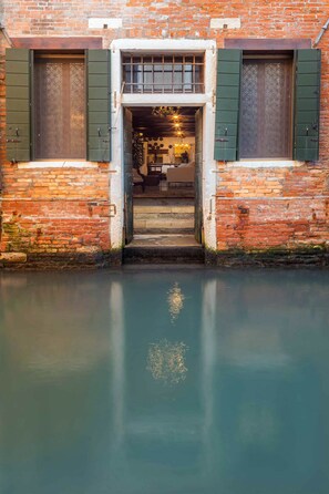 A brick building with a waterway in front, a door with green shutters, and a staircase leading inside. - Unconventional loft-style apartment in San Barnaba (Venecia)