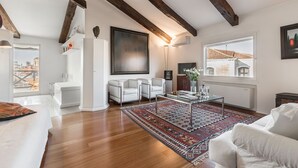 Ein geräumiges Wohnzimmer mit Holzbalken, einem großen Gemälde, einer Couch, einem Couchtisch und Blick auf Gebäude durch ein Fenster.