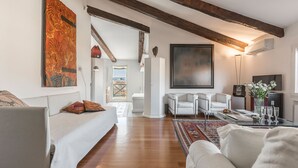 Ein geräumiges Wohnzimmer mit einer Couch, Sesseln und einem Couchtisch. An der Wand hängt ein großes Gemälde und durch eine Tür ist ein Ausblick ins Freie zu sehen.