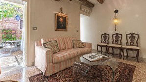 Un salon chaleureux avec un canapé à rayures, une table basse en verre et un tapis à motifs. On aperçoit un coin repas avec une table et des chaises à travers une porte ouverte.