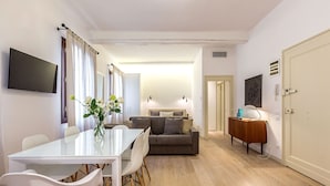 A modern living room with a sofa, a dining table with chairs, a TV mounted on the wall, and a door leading to another room. - Spacious and Charming Apartment Near Fenice Theatre (Venecia)