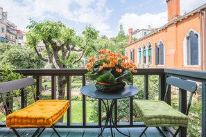 Ein Balkon mit zwei gepolsterten Stühlen und einem Tisch, einer Pflanze mit orangefarbenen Blüten und Blick auf ein Gebäude mit Rundbogenfenstern.