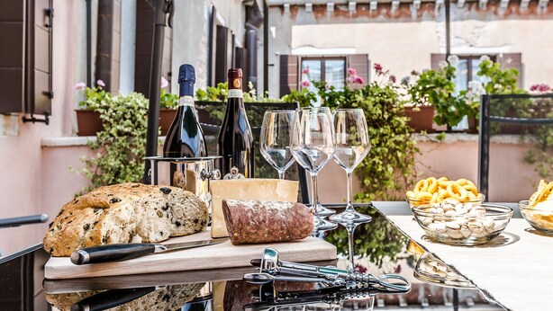A table set with a loaf of bread, cheese, a knife, a bottle, and glasses, with a view of a building and plants in the background.
