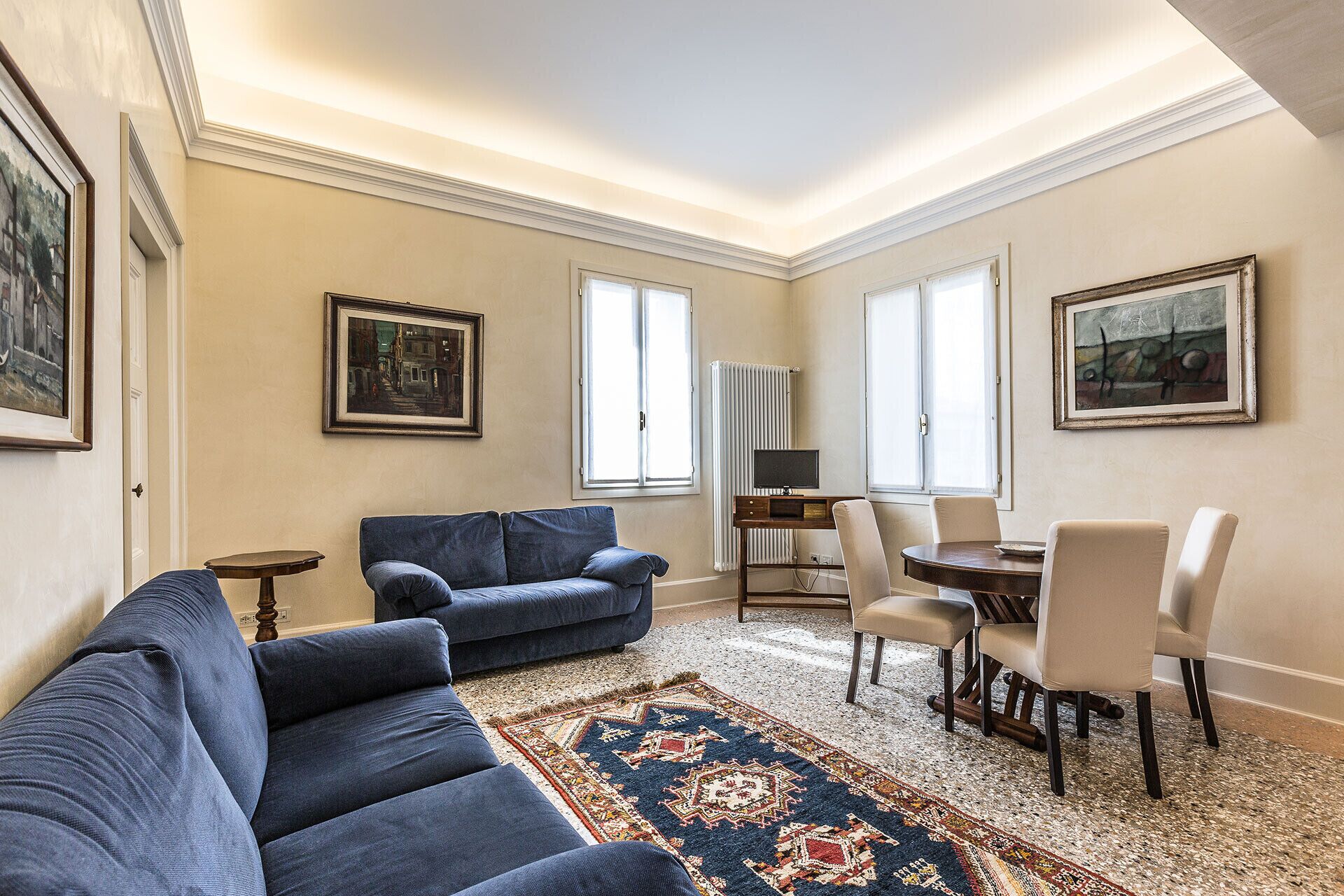 Ein Wohnzimmer mit einem blauen Sofa, einem Essbereich mit Tisch und Stühlen sowie zwei Fenstern mit weißen Vorhängen.