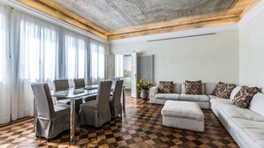 A modern living room with a checkered wooden floor, a glass dining table with chairs, a white sofa with patterned cushions, and a large vase with yellow flowers.