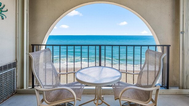 Ein Balkon mit zwei Korbstühlen und einem kleinen runden Tisch, mit Blick auf einen Strand, das Meer und einen wolkenlosen Himmel.