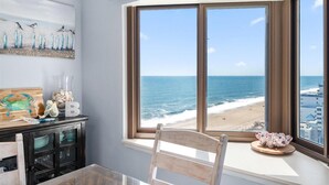 Ein Zimmer mit einem großen Erkerfenster mit Blick auf einen Strand, einem Holztisch mit Dekoration und einem Gemälde an der Wand.