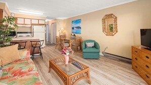 A living room with a sofa, coffee table, dining table, chairs, a TV, and a kitchen area with a refrigerator and stove.