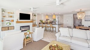 Una sala de estar moderna con chimenea, sofás blancos, una mesa de centro y una cocina al fondo.